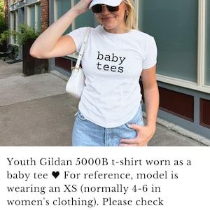 May include: White t-shirt with the text "baby tees" in black, paired with light-wash denim jeans. The model is wearing a white baseball cap and sunglasses. A white purse is worn over the shoulder.