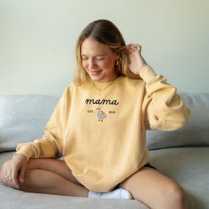 May include: Yellow sweatshirt with the word "mama" in a script font. Below the word "mama" is the text "EST. 2025" and a cartoon goose with a yellow chick. The sweatshirt is a solid yellow color.