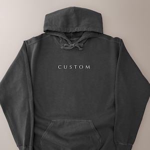 K&ouml;nnte beinhalten: Ein anthrazitfarbener Kapuzenpullover mit Kordelzugkapuze und Vordertasche. Das Wort "CUSTOM" ist in Wei&szlig; auf die Brust gestickt. Der Hoodie ist aus einem weichen, bequemen Material gefertigt.
