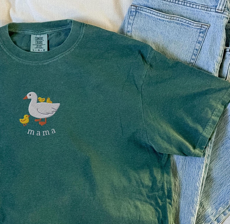 May include: A dark green t-shirt with an embroidered image of a goose and three ducklings. The text "mama" is embroidered below the image.