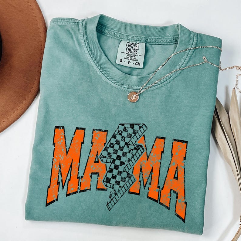 K&ouml;nnte beinhalten: Ein blaugr&uuml;nes T-Shirt mit einer abgenutzten orangefarbenen Grafik, auf der "MAMA" mit einem karierten Blitz in der Mitte steht.