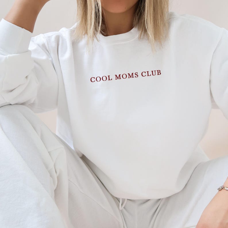 May include: A white sweatshirt with the text "COOL MOMS CLUB" in burgundy lettering. The sweatshirt is part of a matching set with white tracksuit bottoms. The outfit is casual and comfortable.