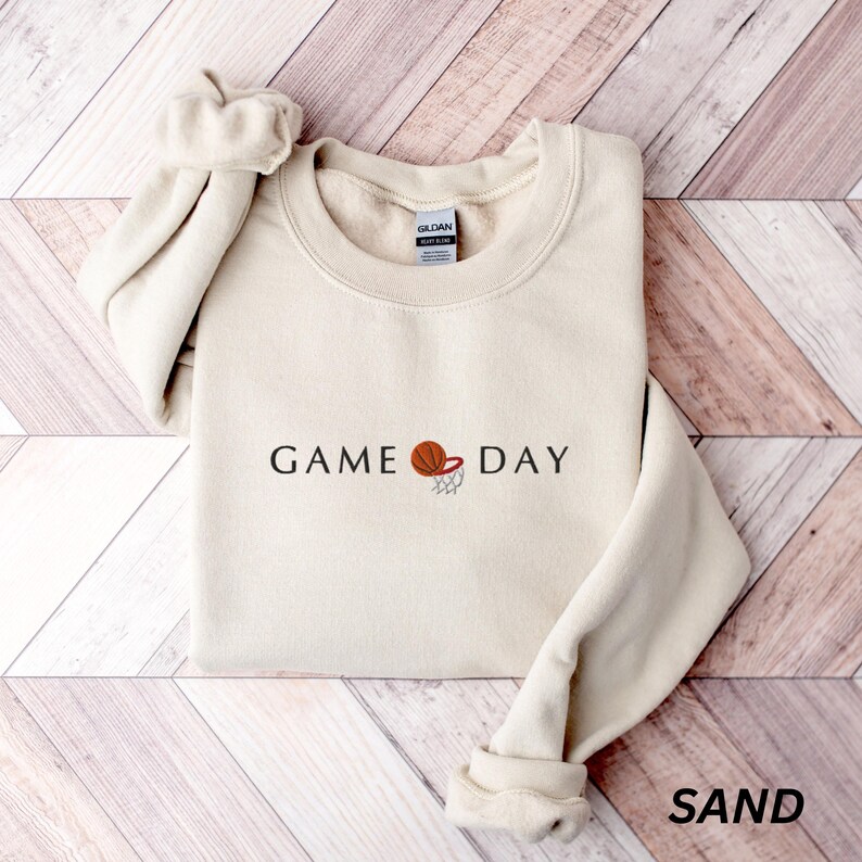 May include: A sand-colored sweatshirt with the words "GAME DAY" and a basketball graphic. The sweatshirt has a crew neck and long sleeves. The word "SAND" is printed in the bottom right corner.