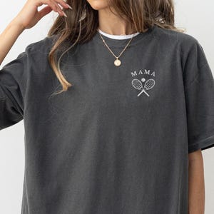 May include: A dark grey t-shirt with the word "MAMA" and a tennis racket graphic printed on the front.