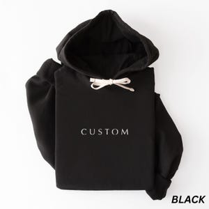 May include: Black hoodie with a drawstring hood and the word "CUSTOM" printed in white. The word "BLACK" is printed in white at the bottom right corner. The hoodie is made of a soft material.