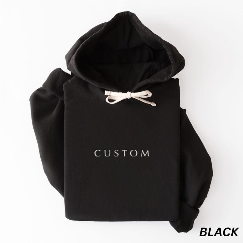 May include: Black hoodie with a drawstring hood and the word "CUSTOM" printed in white. The word "BLACK" is printed in white at the bottom right corner. The hoodie is made of a soft material.