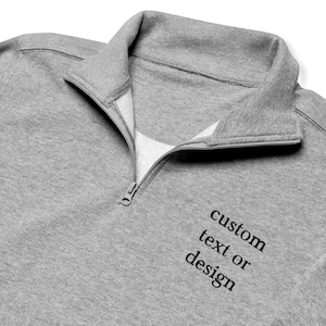 May include: A gray quarter-zip sweatshirt with a folded collar and a partially open zipper. The text "custom text or design" is printed in black on the lower right side of the sweatshirt. The sweatshirt is made of a soft, comfortable material.