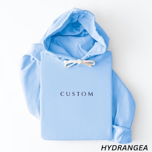 May include: A light blue hoodie with a drawstring hood and long sleeves. The word "CUSTOM" is printed in dark blue on the front. The word "HYDRANGEA" is printed in black at the bottom.