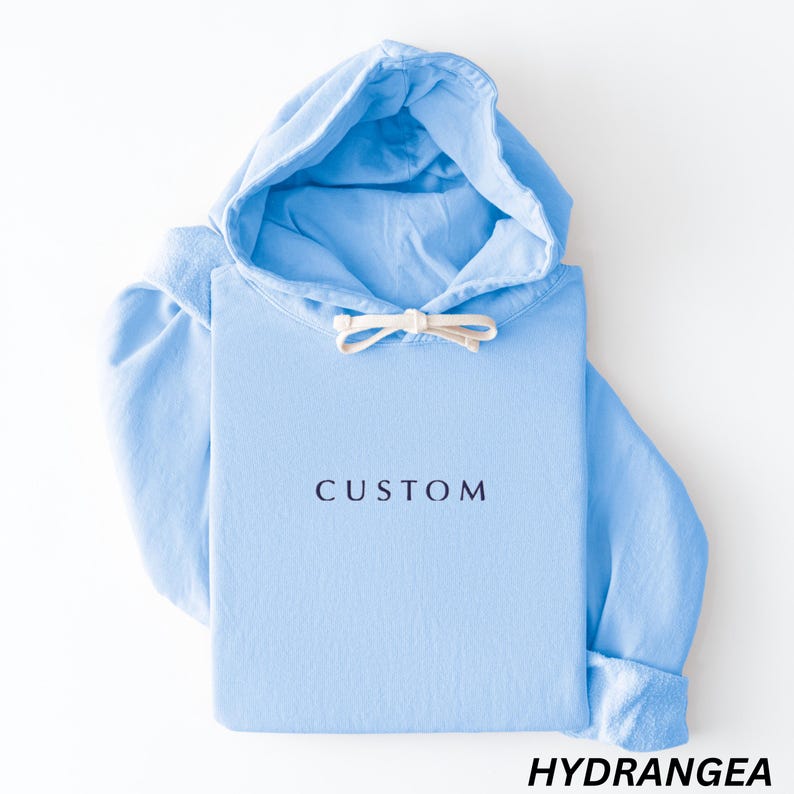 May include: A light blue hoodie with a drawstring hood and long sleeves. The word "CUSTOM" is printed in dark blue on the front. The word "HYDRANGEA" is printed in black at the bottom.