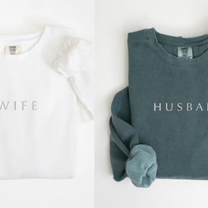 May include: Two long-sleeved shirts, one white with the word "WIFE" and one teal with the word "HUSBAND" printed on them.
