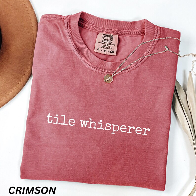 K&ouml;nnte beinhalten: Karmesinrotes T-Shirt mit dem wei&szlig;en Text "tile whisperer". Das Shirt hat einen Rundhalsausschnitt und kurze &Auml;rmel. Eine silberne Halskette mit rundem Anh&auml;nger liegt auf dem Shirt. Das Shirt ist von Comfort Colors.