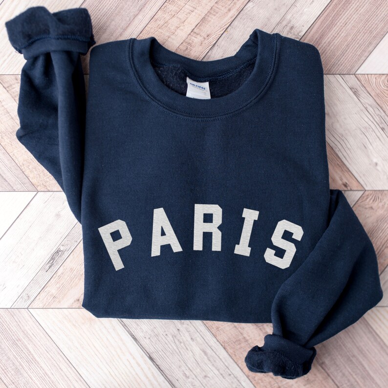 Puede incluir: Una sudadera azul marino con la palabra "PARIS" impresa en blanco en la parte delantera.