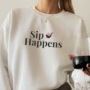 May include: White sweatshirt with the text "Sip Happens" and a graphic of a red wine glass. A person is holding a glass of red wine.