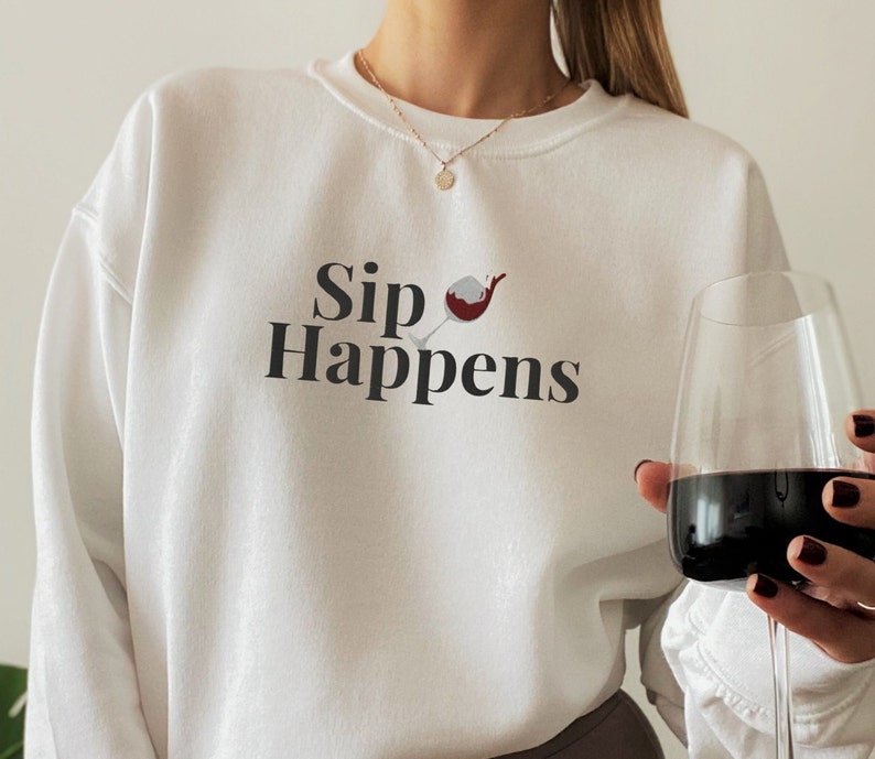 May include: White sweatshirt with the text "Sip Happens" and a graphic of a red wine glass. A person is holding a glass of red wine.