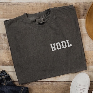 May include: A dark gray t-shirt with the word "HODL" printed in white on the front. The shirt is made of a soft, comfortable material and has a relaxed fit.