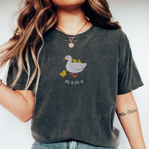 May include: A dark gray t-shirt with a white embroidered design of a mama duck with three ducklings. The text "mama" is embroidered below the duck.