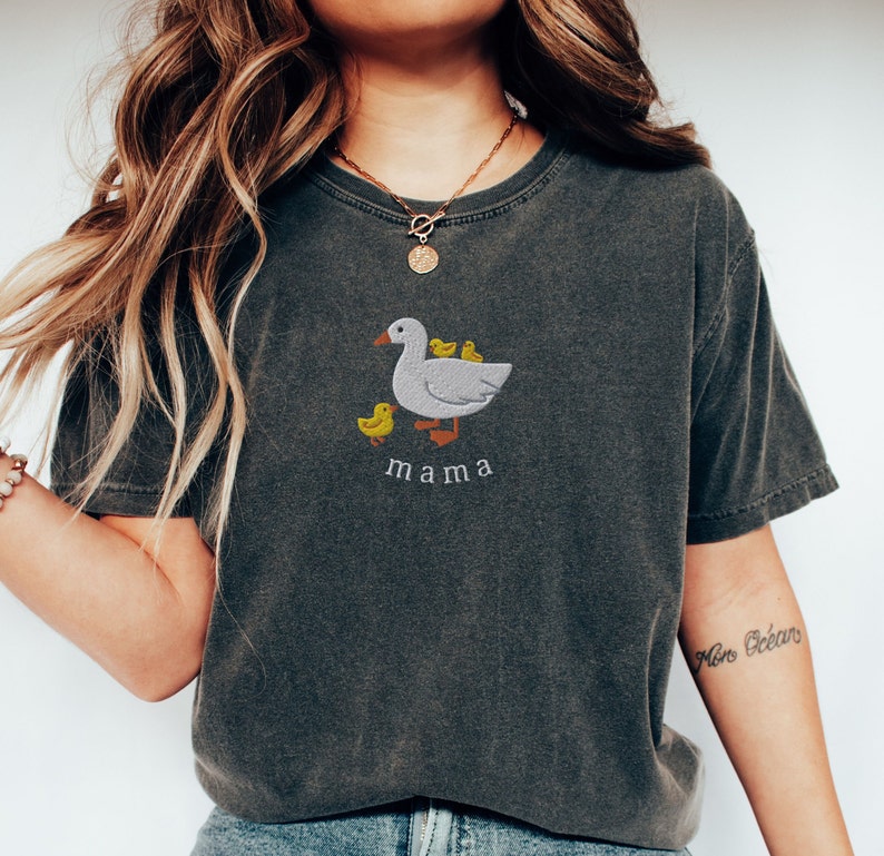 May include: A dark gray t-shirt with a white embroidered design of a mama duck with three ducklings. The text "mama" is embroidered below the duck.