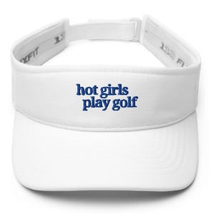 May include: White visor with the embroidered text "hot girls play golf" in blue. The visor has a curved brim and an adjustable strap. The text is in a playful, casual font.