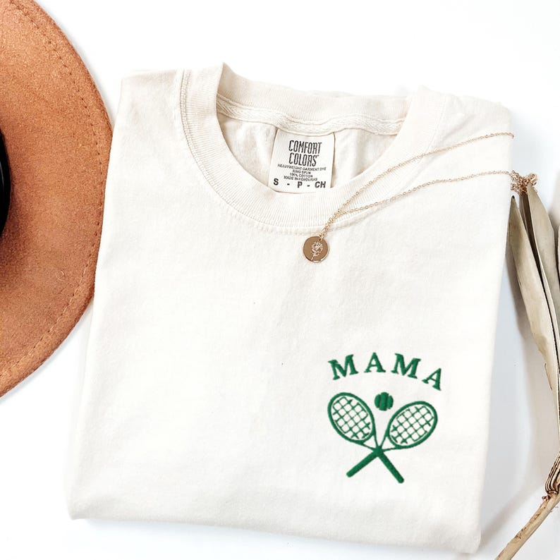 May include: A white t-shirt with green embroidery of two tennis rackets crossed with a tennis ball in the middle. The text "MAMA" is above the rackets.