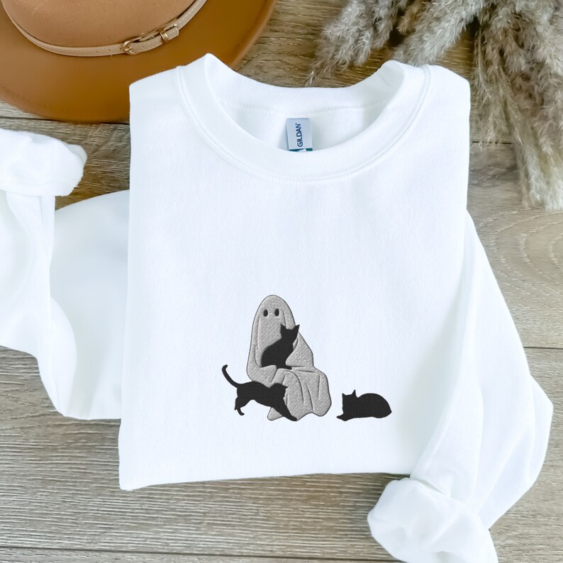 K&ouml;nnte beinhalten: Wei&szlig;er Pullover mit einem grauen Geist und drei schwarzen Katzen, die auf der Vorderseite gestickt sind. Der Geist h&auml;lt eine der Katzen.