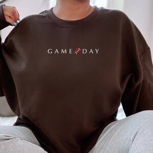 Może przedstawiać: Ciemnobrązowa bluza z białym napisem "GAME DAY" i grafiką piłki nożnej. Top z długim rękawem jest w jednolitym kolorze. Temat: odzież sportowa na dzień meczu.