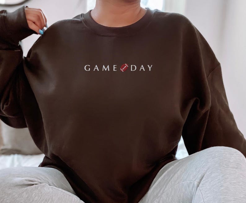 Może przedstawiać: Ciemnobrązowa bluza z białym napisem "GAME DAY" i grafiką piłki nożnej. Top z długim rękawem jest w jednolitym kolorze. Temat: odzież sportowa na dzień meczu.
