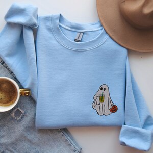 May include: Light blue sweatshirt with a white ghost holding a pumpkin and a beverage. The ghost has two eyes and a mouth.