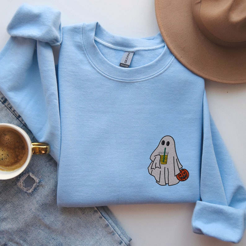 May include: Light blue sweatshirt with a white ghost holding a pumpkin and a beverage. The ghost has two eyes and a mouth.