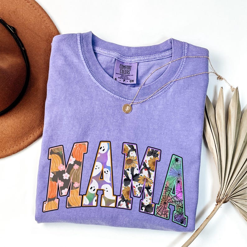Puede incluir: Camiseta morada con la palabra "MAMA" en letras grandes y coloridas. Cada letra est&aacute; llena de un patr&oacute;n diferente con tem&aacute;tica de Halloween, que incluye calabazas, fantasmas, ara&ntilde;as y girasoles.