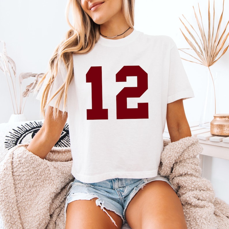 May include: White t-shirt with the number 12 in a bold, maroon font. The shirt is short-sleeved and appears to be made of a soft material. The t-shirt is worn with denim shorts.