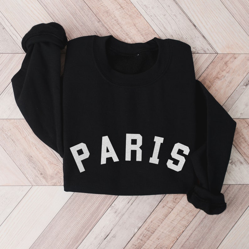 Puede incluir: Sudadera negra con texto blanco que dice "PARIS" en una fuente en negrita y en bloque.