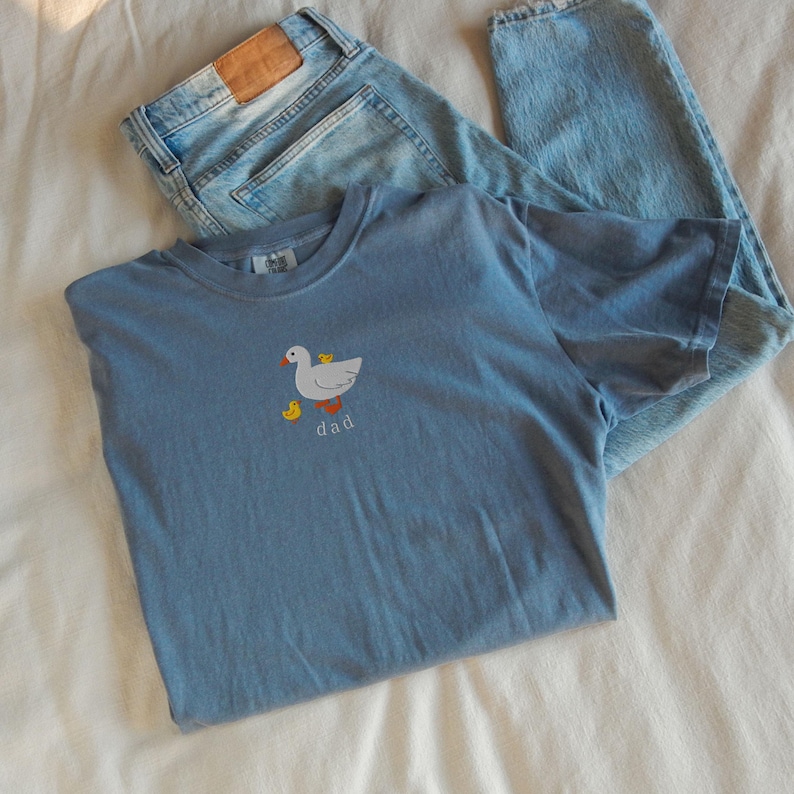 May include: A light blue t-shirt with a duck and duckling graphic and the word "dad" in red. The shirt is paired with light wash blue jeans with a leather patch.
