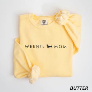 Puede incluir: Sudadera amarilla clara con las palabras "WEENIE MOM" y una silueta negra de perro salchicha. La palabra "BUTTER" est&aacute; impresa en la parte inferior. Las mangas est&aacute;n anudadas. Prenda c&oacute;moda e informal.