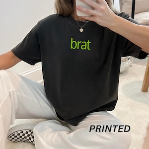 May include: A black t-shirt with a green "brat" graphic printed on the front. The shirt is being worn by a person with white pants.