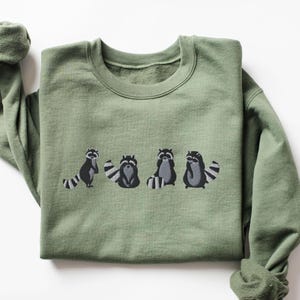 May include: A green sweatshirt with a graphic of four raccoons in a line. The raccoons are black and gray with white accents.