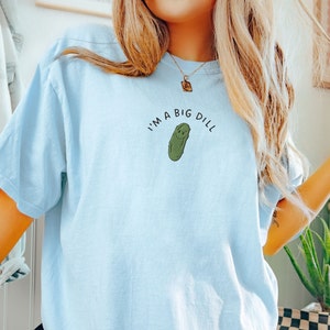 Op de afbeelding: Een lichtblauw T-shirt met de tekst "I'm a big dill" en een cartoon-komkommer met een glimlach.