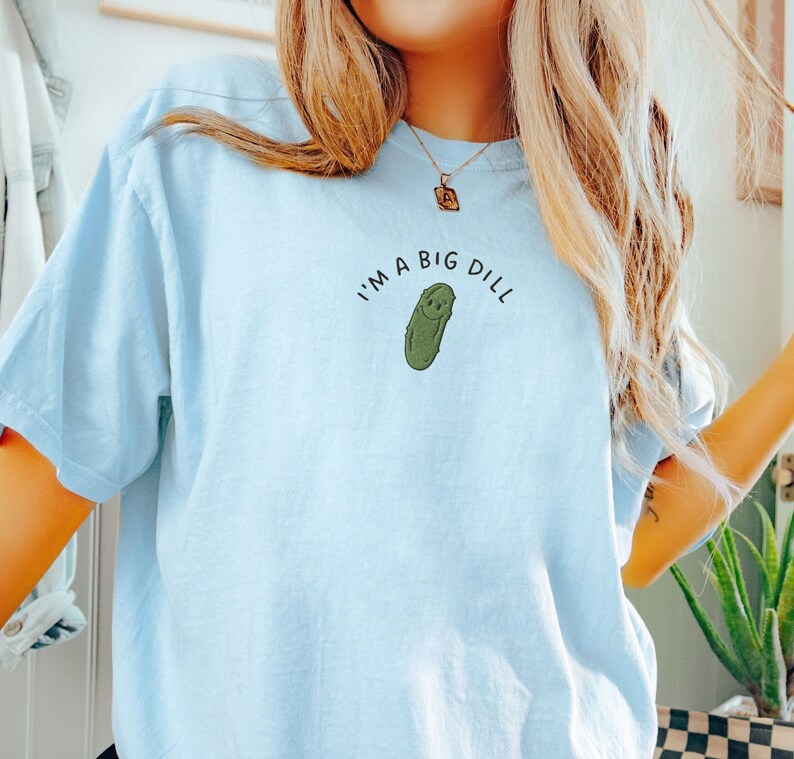Op de afbeelding: Een lichtblauw T-shirt met de tekst "I'm a big dill" en een cartoon-komkommer met een glimlach.