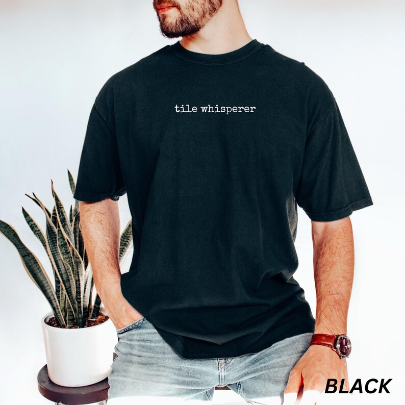 K&ouml;nnte beinhalten: Schwarzes T-Shirt mit dem wei&szlig;en Text "tile whisperer". Das Shirt hat eine dunkle Farbe und eine lockere Passform. Das Wort "BLACK" ist in Wei&szlig; in der unteren rechten Ecke des Bildes aufgedruckt.
