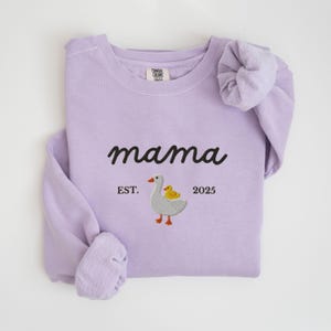 May include: A light purple sweatshirt with the word "mama" in black script. Below the word "mama" is an embroidered design of a goose and a duckling, with the text "EST. 2025". The sleeves are folded.