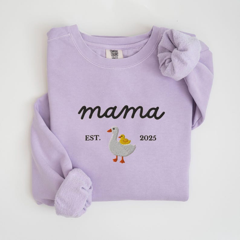May include: A light purple sweatshirt with the word "mama" in black script. Below the word "mama" is an embroidered design of a goose and a duckling, with the text "EST. 2025". The sleeves are folded.