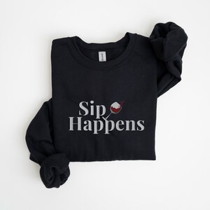 May include: Black sweatshirt with a white graphic that says "Sip Happens" and a red wine glass with a white rim.