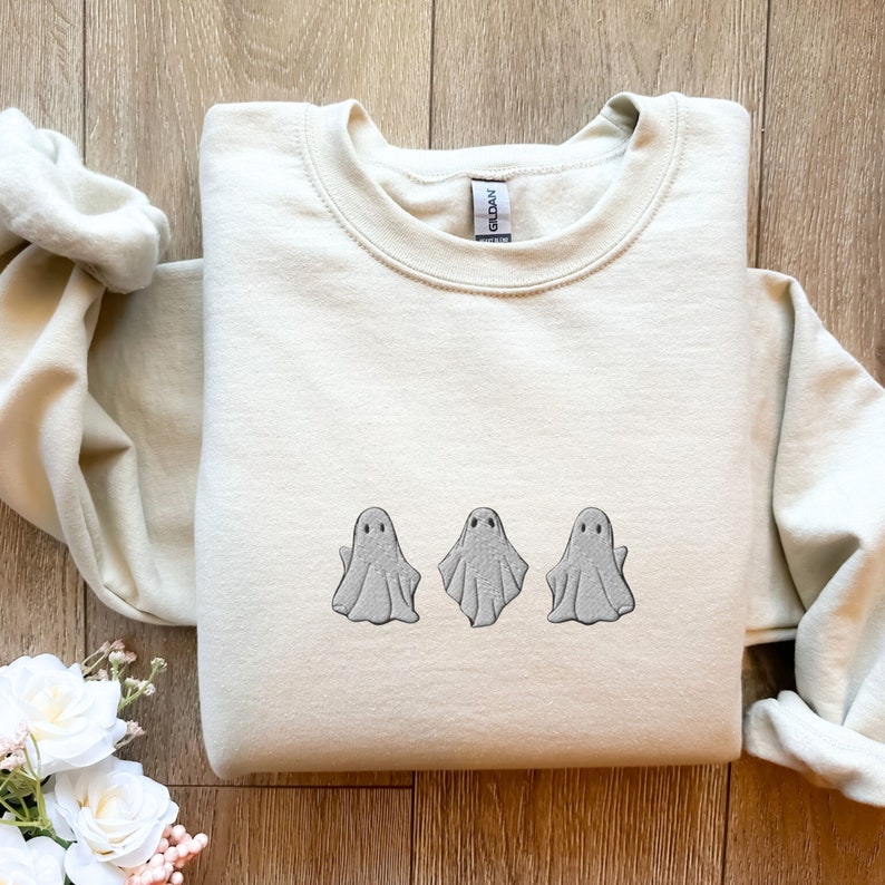 May include: A cream-colored sweatshirt with three embroidered ghosts in a row on the front. The ghosts are gray and have simple facial features.