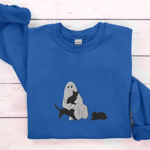 K&ouml;nnte beinhalten: Ein k&ouml;nigsblauer Pullover mit einem wei&szlig;en Geist und drei schwarzen Katzen, die auf der Vorderseite gestickt sind. Der Geist h&auml;lt eine der Katzen in seinen Armen.