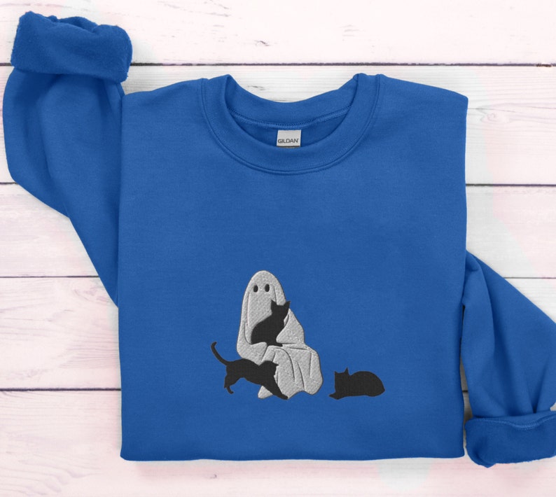 K&ouml;nnte beinhalten: Ein k&ouml;nigsblauer Pullover mit einem wei&szlig;en Geist und drei schwarzen Katzen, die auf der Vorderseite gestickt sind. Der Geist h&auml;lt eine der Katzen in seinen Armen.