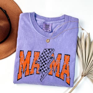K&ouml;nnte beinhalten: Ein lila T-Shirt mit einer abgenutzten orangefarbenen Grafik, auf der "MAMA" mit einem karierten Blitz in der Mitte steht. Das Shirt ist gefaltet und auf einer wei&szlig;en Oberfl&auml;che abgelegt.