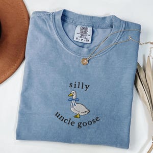 May include: A light blue Comfort Colors t-shirt with the embroidered phrase "silly uncle goose" and a cartoon goose wearing a blue bow tie. A gold necklace with a pendant is draped across the shirt.