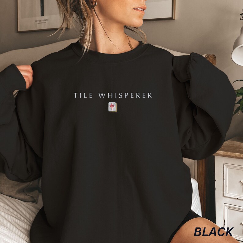 May include: Black sweatshirt with the text "TILE WHISPERER" in white, above a small, square graphic. The graphic is a red and white tile symbol. The word "BLACK" is printed in white at the bottom right.