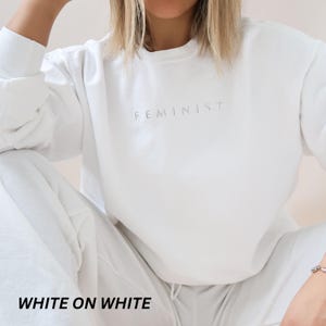 Pu&ograve; includere: Un completo bianco di felpa e pantaloni della tuta abbinati. La parola "FEMINIST" &egrave; stampata in modo sottile sul petto della felpa. La frase "WHITE ON WHITE" &egrave; stampata in grassetto, in nero nella parte inferiore dell'immagine.
