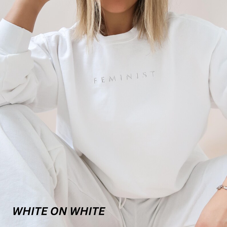 Pu&ograve; includere: Un completo bianco di felpa e pantaloni della tuta abbinati. La parola "FEMINIST" &egrave; stampata in modo sottile sul petto della felpa. La frase "WHITE ON WHITE" &egrave; stampata in grassetto, in nero nella parte inferiore dell'immagine.