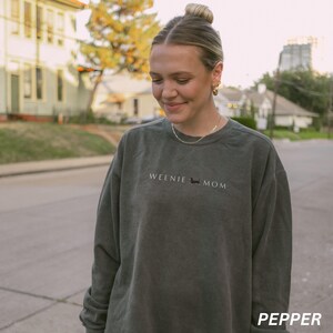 Puede incluir: Una sudadera verde oscuro con el texto "WEENIE MOM" y una ilustraci&oacute;n de perro salchicha. La palabra "PEPPER" est&aacute; en la esquina inferior derecha. La sudadera la lleva una persona en la calle.